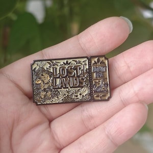 May include: A rectangular metal enamel pin designed as a vintage ticket with a dark bronze finish. The pin features the text LOST LANDS and ADMIT ONE, with engraved illustrations of a dinosaur, a volcano, and a small creature carrying a backpack.