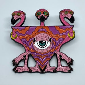 May include: A pink enamel pin featuring three pink flamingos with sunglasses, forming a triangle shape with a large eye in the center. The eye is surrounded by a purple and orange wavy pattern.