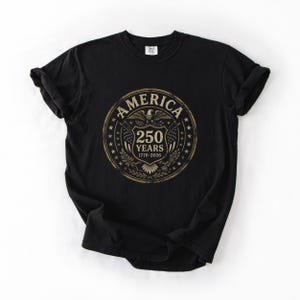 May include: Black t-shirt featuring a circular design with the word "AMERICA" at the top. The design includes an eagle, stars, and the text "250 YEARS 1776-2026". The shirt is made of a soft material and has a classic crew neck.