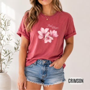 May include: Crimson-colored t-shirt featuring a watercolor floral design with the text "Petal Whisper." The shirt has rolled-up sleeves and is paired with denim shorts. The word "CRIMSON" is displayed at the bottom right.