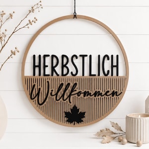 Könnte beinhalten: Ein rundes Wandschild aus Holz mit dem schwarzen Schriftzug HERBSTLICH Willkommen und einer schwarzen Ahornblatt-Silhouette. Das Schild hat einen hellbraunen Holzrahmen mit vertikalen Lamellen und hängt an einer dünnen schwarzen Kordel.