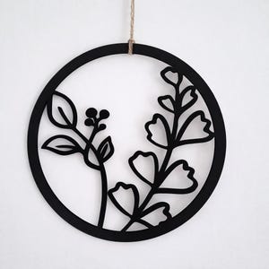 Könnte beinhalten: Schwarze kreisförmige Wandkunst mit einem botanischen Design. Das Kunstwerk zeigt stilisierte Blätter und Beeren in einem kreisförmigen Rahmen. Eine natürliche Schnurschlaufe ist zum Aufhängen angebracht. Das Design ist modern und minimalistisch.