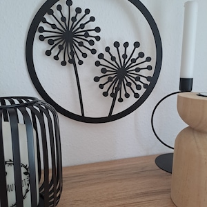 Könnte beinhalten: Ein schwarzes Metall-Wandkunstwerk mit Löwenzahnmotiven hängt an einer weißen Wand. Die Kunst zeigt zwei Löwenzahnsamenköpfe in einem kreisförmigen Rahmen. Ein schwarzer Kerzenhalter mit einer brennenden Kerze und ein Kerzenhalter aus Holz befinden sich auf einer Holzoberfläche.