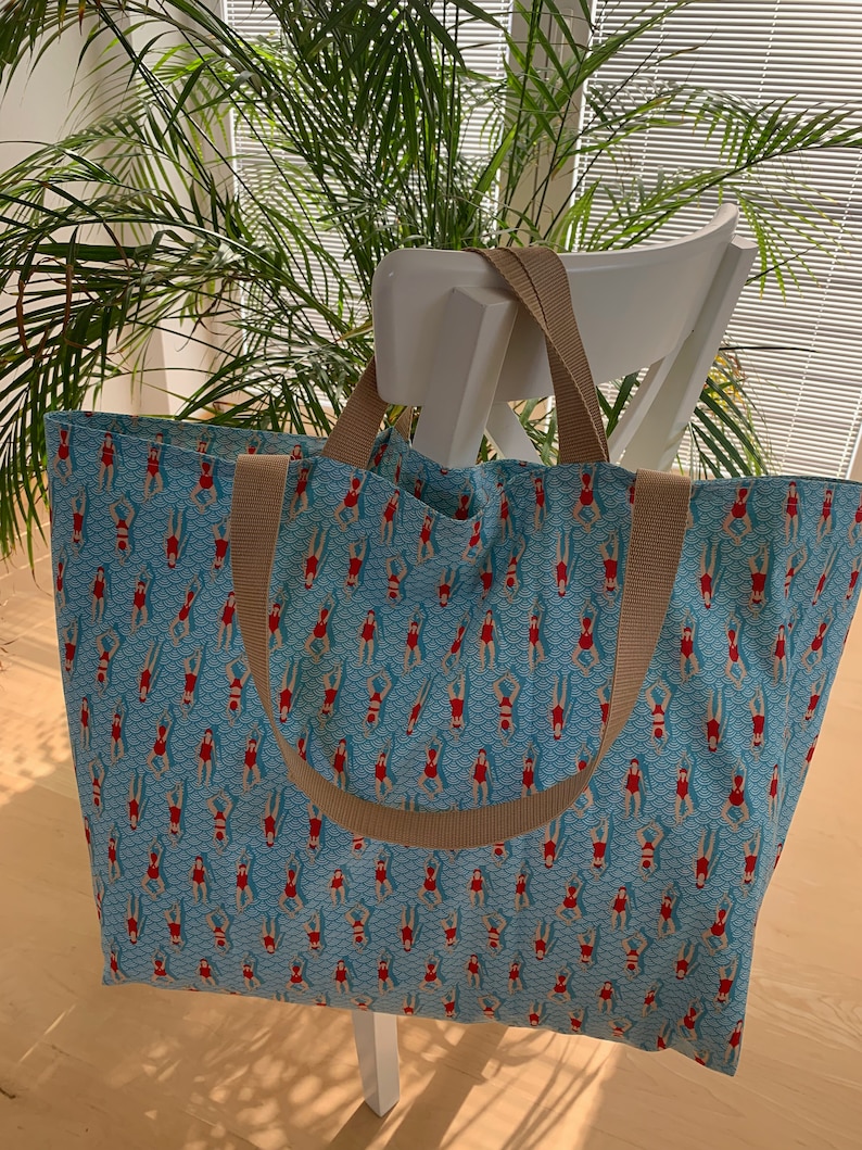 May include: A large blue tote bag with brown straps. The bag has a repeating pattern of red figures in swimsuits floating on a blue background.