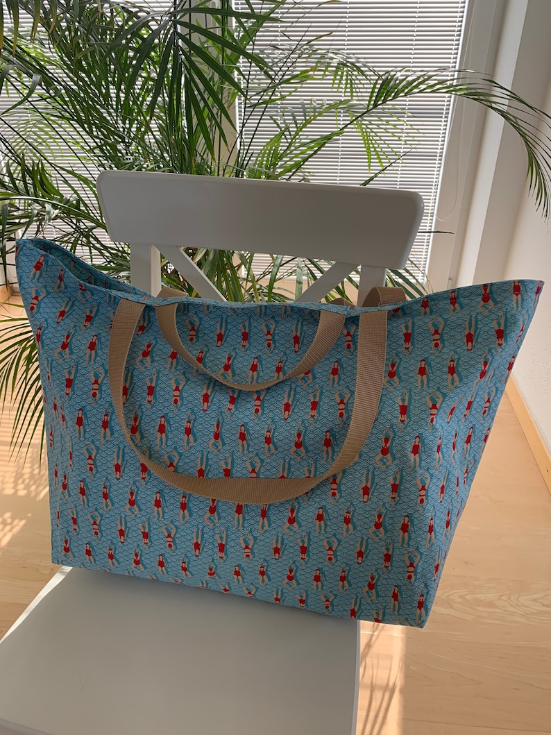 May include: A large blue tote bag with tan straps. The bag is printed with a repeating pattern of women in red swimsuits swimming in a blue pool.