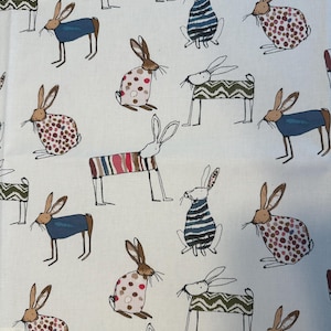 Könnte beinhalten: Cremefarbener Stoff mit einem sich wiederholenden Muster aus skurrilen Hasen. Die Hasen sind in verschiedenen Posen und Outfits dargestellt, darunter Pullover und gemusterte Mäntel. Die Farbpalette umfasst braune, blaue, grüne und rote Akzente.