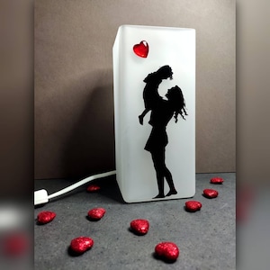 Può includere: Una lampada in vetro bianco smerigliato con una silhouette di una persona che tiene un bambino. La lampada ha un cuore rosso in cima ed è accesa. Ci sono cuori di glitter rossi sparsi intorno alla lampada.