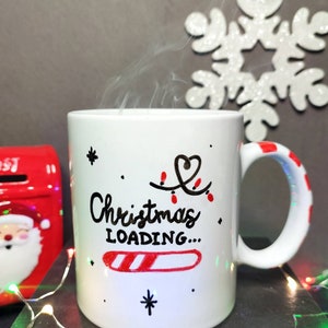 May include: White ceramic mug with a black and red design. The mug has a black handwritten message that says "Christmas Loading..." with a candy cane progress bar.