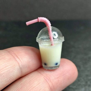 Op de afbeelding: Miniatuur bubble tea drankje in een doorzichtig plastic bekertje met een roze rietje en deksel. De drank bevat een romige, gebroken witte vloeistof en donkerblauwe tapiocaparels. Het roze rietje is gebogen.