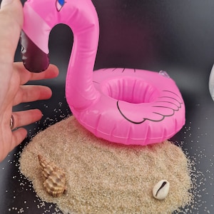 以下が含まれることがあります： 黒と白の目のデザインが施されたピンクのインフレータブルフラミンゴドリンクホルダー。フラミンゴは砂の山に2つの貝殻と一緒に座っています。