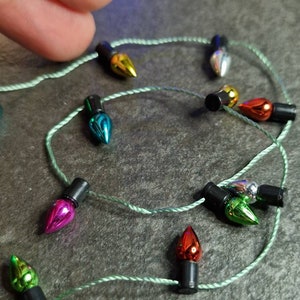 May include: A close-up of a string of miniature Christmas lights. The bulbs are in various colours, including gold, silver, red, green, and blue, and are connected by a thin, green wire. The lights are set against a dark grey background.