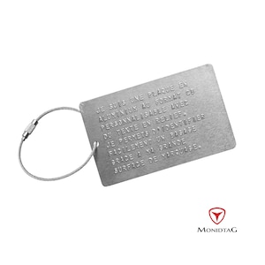 Peut inclure: Une étiquette à bagages en argent avec un anneau en métal et une chaîne en câble. L'étiquette porte un message gravé qui dit : "JE SUIS UNE PLAQUE EN ALUMINIUM AU FORMAT CB PERSONNALISABLE AVEC DU TEXTE EN RELIEF. JE PERMETS D'IDENTIFIER FACILEMENT UN BAGAGE GRACE A MA GRANDE SURFACE DE MARQUAGE." L'étiquette porte également le mot "MONIDTAG" imprimé dessus.