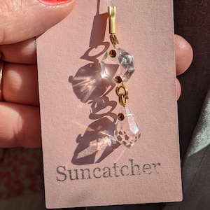 Könnte beinhalten: Ein klarer Kristall-Sonnenfänger mit goldfarbenen Akzenten hängt an einem rosa Anhänger. Der Sonnenfänger hat mehrere facettierte Kristallperlen und einen tropfenförmigen Kristall am unteren Ende. Auf dem Anhänger steht "Suncatcher".