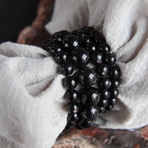 Puede incluir: Primer plano de un brazalete de cuentas negras con múltiples hileras de cuentas redondas brillantes. El brazalete está apoyado sobre un trozo de tela blanca.
