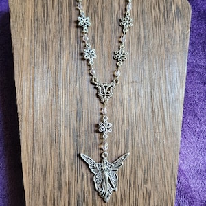 May include: A silver-toned necklace featuring a series of small metal flower charms and clear faceted beads. The chain leads to an ornate filigree connector piece, followed by a single flower charm and a large fairy pendant with spread wings.