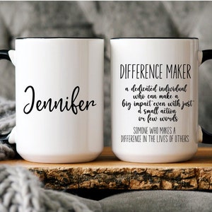May include: Two white ceramic mugs with black handles and black rims. One mug has the name "Jennifer" written on it. The other mug has the text "DIFFERENCE MAKER" and a definition of the term.