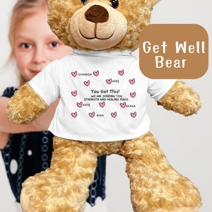 May include: A light brown teddy bear wearing a white t-shirt with the text "You Got This! We are sending you strength and healing vibes."  The shirt has pink hearts with names written inside each heart. The bear is sitting on a white surface.  Get Well Bear.