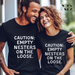 May include: A couple wearing black t-shirts with the text "CAUTION: EMPTY NESTERS ON THE LOOSE." printed in white. They are smiling and looking at each other.