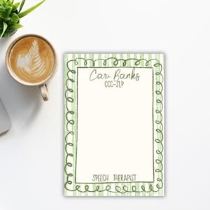 May include: A notepad with a green striped border and the text "Car Banks CCC-SLP SPEECH THERAPIST" in a handwritten style. The notepad is on a white surface, next to a cup of coffee and a small plant.