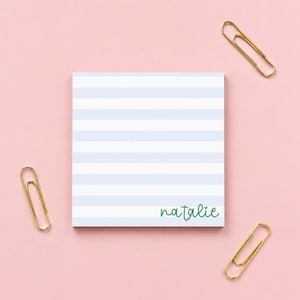 May include: A square notepad featuring horizontal light blue and white stripes with the name "natalie" printed in green cursive script at the bottom right corner, accompanied by three gold-toned metal paper clips positioned around the notepad.