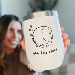 May include: White insulated tumbler with a silver rim, featuring a hand-drawn clock design and the phrase "off the clock" in black. The tumbler is designed to keep beverages at the desired temperature.