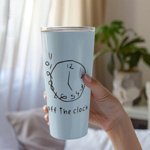 May include: A light blue tumbler with a black hand-drawn clock design and the words "off the clock." The tumbler has a silver lid and is held by a person. The design features the numbers 12, 2, 3, 4, 5, and 11.