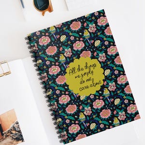 May include: A spiral-bound notebook with a floral pattern of pink flowers, green leaves, and yellow birds on a black background. The notebook has the text "All the things we simply do not care about" on a yellow background.