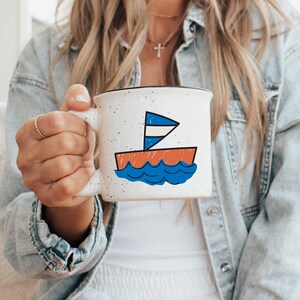 May include: White ceramic mug with a black rim and a sailboat illustration. The sailboat features a blue and white sail, an orange hull, and blue waves. The mug has a speckled design.