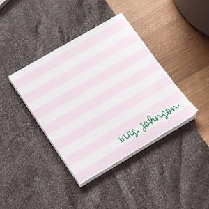 May include: A square notepad with pink and white horizontal stripes. The words "mrs. johnson" are written in green script in the lower right corner. The notepad is on a gray surface.