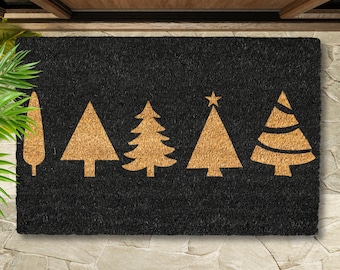 May include: Black rectangular doormat with a series of five gold-colored Christmas tree designs. The trees vary in shape and style, with one featuring a star at the top. The mat is made of a textured material.