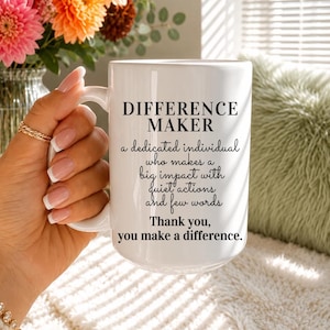 May include: A white ceramic mug featuring the black text: DIFFERENCE MAKER, a dedicated individual who makes a big impact with quiet actions and few words. Thank you, you make a difference. The mug has a smooth finish and a curved handle.