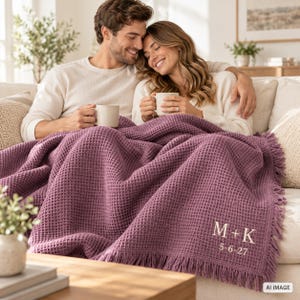 Pode incluir: Uma manta de malha waffle roxo-escura com bordas de franjas e bordado personalizado em fio branco que diz M + K seguido pela data 5-6-27. A manta está sobre um sofá, exibindo o seu padrão de grade texturizado.
