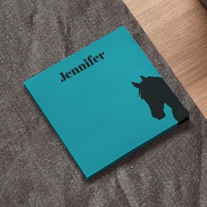 May include: A teal square notepad with the name "Jennifer" printed in black. A black silhouette of a horse's head is on the right side of the notepad. The notepad is on a gray textured surface.