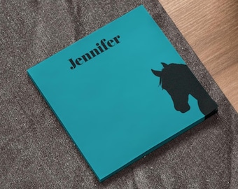 May include: A teal square notepad with the name "Jennifer" printed in black. A black silhouette of a horse's head is on the right side of the notepad. The notepad is on a gray textured surface.