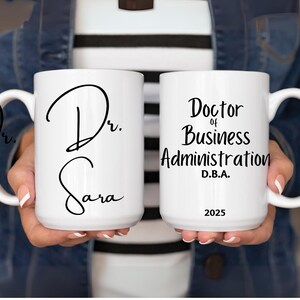 May include: Two white ceramic mugs. One mug has the text "Dr. Sara" in black script. The other mug has the text "Doctor of Business Administration D.B.A. 2025" in black. Both mugs have a white handle.