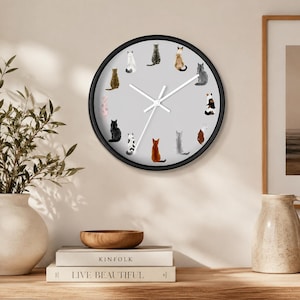 May include: A round wall clock with a black frame and a light gray face featuring twelve illustrations of cats in various colors and patterns arranged in a circle. The clock has two white hands and a thin white second hand at the center.