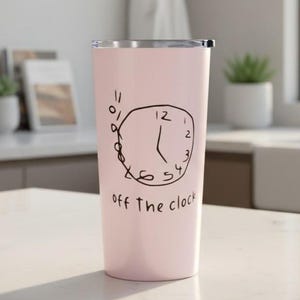 May include: A light pink tumbler with a silver rim. The tumbler features a hand-drawn clock design in black ink, with the words "off the clock" below the clock. The tumbler is cylindrical and appears to be made of stainless steel.
