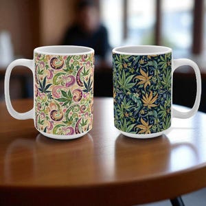 Può includere: Due tazze in ceramica bianca con motivi a foglie di cannabis. Una tazza presenta un design paisley con foglie verdi, viola e gialle su sfondo crema. L'altra ha foglie verdi e dorate su sfondo blu scuro.
