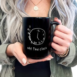 May include: Black ceramic mug with a white hand-drawn clock design and the words "off the clock" in a handwritten font. The mug has a large handle and is held by a person.