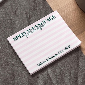 May include: A square notepad featuring horizontal light pink and white stripes. The text SPEECH-LANGUAGE pathologist is printed in dark green at the top, and the name Olivia Johnson, CCC-SLP is printed in dark green at the bottom right corner.