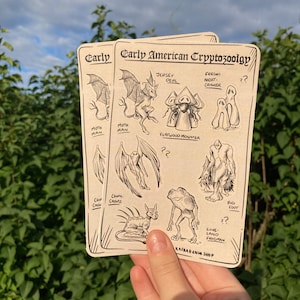 Puede incluir: Dos tarjetas color crema con ilustraciones de críptidos, incluyendo Mothman, Jersey Devil y Bigfoot. Las tarjetas están etiquetadas como "Early American Cryptozoology" con el nombre de la tienda "Kafkaesque Shop" en la parte inferior. El fondo es un arbusto verde y un cielo nublado.