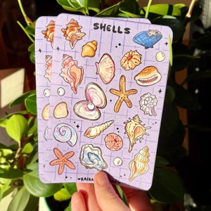 May include: A rectangular sticker sheet with a light purple grid background featuring various illustrated seashells, starfish, and pearls. The text SHELLS is printed at the top in black, and the text KAFKA SHOP is printed at the bottom in black.