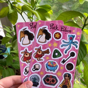 May include: Two pink sticker sheets with various cartoon illustrations, including an angel, a creature playing a trumpet, and a blue octopus. The text "Pink Palace" is visible on the stickers. The stickers are held in front of a background of green leaves.