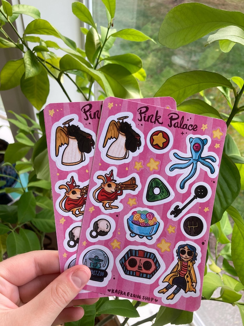 May include: Two pink sticker sheets with various cartoon illustrations, including an angel, a creature playing a trumpet, and a blue octopus. The text "Pink Palace" is visible on the stickers. The stickers are held in front of a background of green leaves.