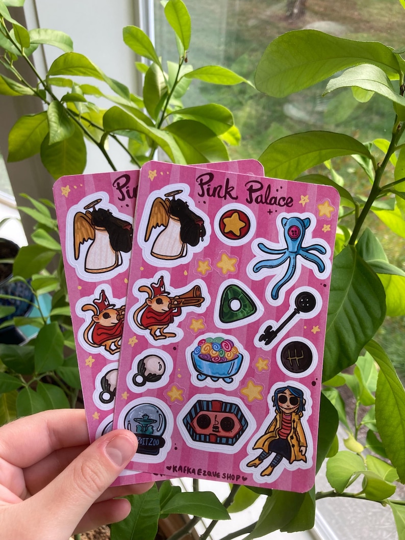 May include: Two pink sticker sheets with various cartoon illustrations, including angels, a key, a blue octopus, and a character in a yellow coat. The text "Pink Palace" and "Kafka Ezque Shop" are visible.