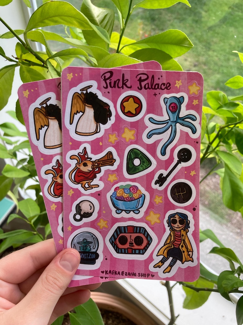 May include: Two pink sticker sheets with various illustrations, including an angel, a star, a key, and a character in a yellow coat. The stickers have a pink background with the text "Pink Palace" and "Kafkaesque Shop".