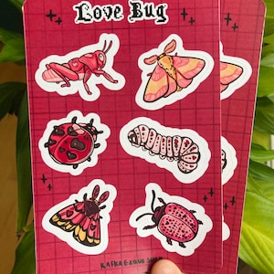 May include: Two red cards with insect stickers. The stickers include a grasshopper, moths, a ladybug, a caterpillar, and a beetle. The text "Love Bug" is at the top, and "Kafka Love Shop" is at the bottom.