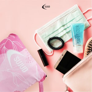 May include: A pink and white patterned drawstring bag with the word "SUJUD" printed on it. The bag is lying on a pink surface with other items, including a face mask, hand sanitizer, a hair tie, lipstick, and a smartphone.