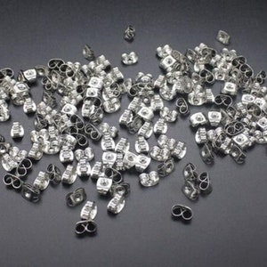 May include: A pile of silver-coloured earring backs scattered on a dark surface. The earring backs are butterfly style, with a post insertion point and a curved clasp. The image shows a close-up view of the earring findings.