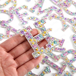 May include: A close-up of a letter "H" covered in iridescent rhinestones, held against a background of other letters. The gems are in shades of pink, purple, yellow, and clear, creating a sparkling effect. These letters are likely for craft projects.
